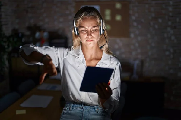 Young blonde woman working at the office at night looking unhappy and angry showing rejection and negative with thumbs down gesture. bad expression.