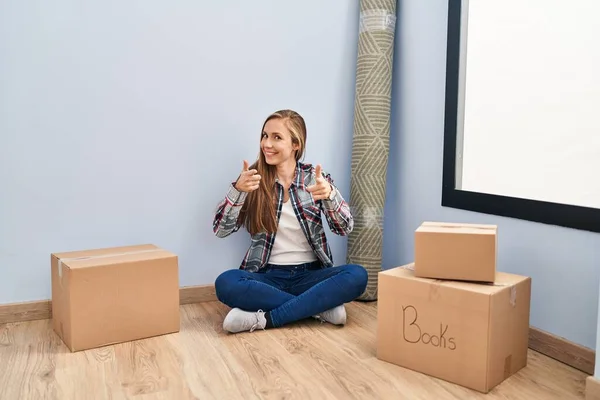 Young blonde woman sitting on the floor moving to a new home pointing fingers to camera with happy and funny face. good energy and vibes.
