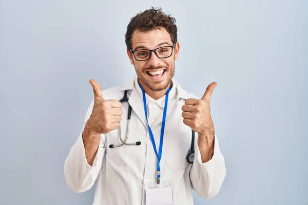医師の制服を着た若いヒスパニック系の男と手で積極的なジェスチャーを行う聴診器の成功サイン 親指を笑顔と幸せ 陽気な表情と勝者のジェスチャー — ストック写真