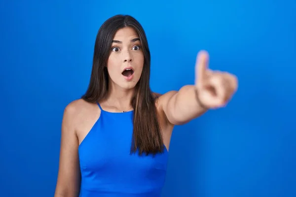 Hispanic woman standing over blue background pointing with finger surprised ahead, open mouth amazed expression, something on the front