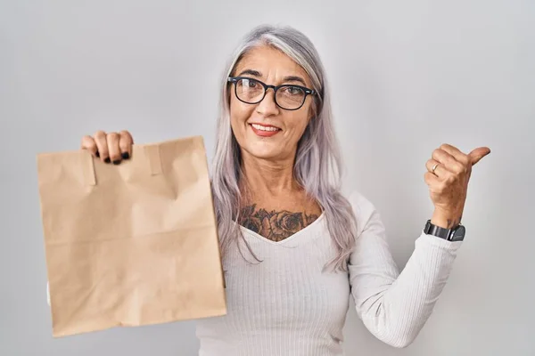 Middle age woman with grey hair holding take away paper bag pointing thumb up to the side smiling happy with open mouth