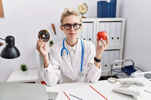 Young Doctor Woman Holding Red Apple Donut Clinic Relaxed Serious — Foto Stock