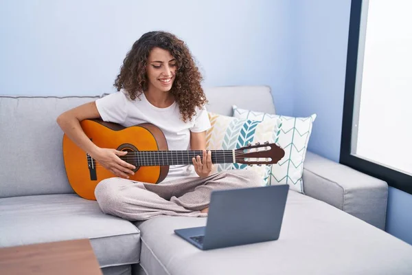 Young beautiful hispanic woman having online classical guitar class sitting on sofa at home