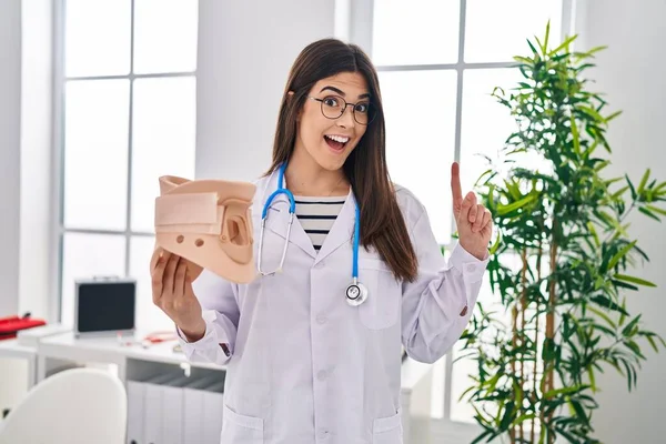 Young brunette doctor woman holding cervical neck collar surprised with an idea or question pointing finger with happy face, number one
