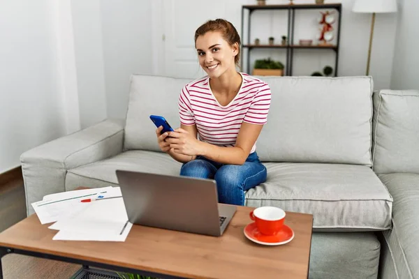 Akıllı telefon ve dizüstü bilgisayar kullanan genç bir kadın evdeki kanepede oturuyor.