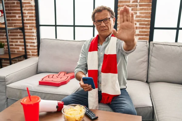 Senior man watching football looking at smartphone with open hand doing stop sign with serious and confident expression, defense gesture