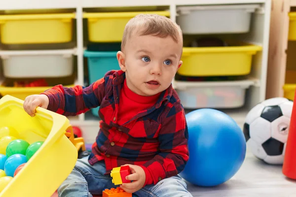 Anaokulunun zemininde oturan, taşaklarıyla oynayan sevimli sarışın çocuk.
