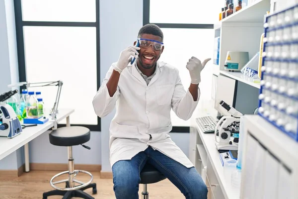 African american man working at scientist laboratory speaking on the phone pointing thumb up to the side smiling happy with open mouth 