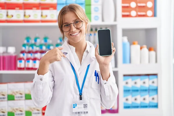 Young caucasian woman working at pharmacy drugstore showing smartphone screen pointing finger to one self smiling happy and proud