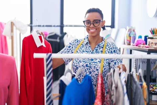 African american woman tailor smiling confident holding clothes on rack at tailor shop
