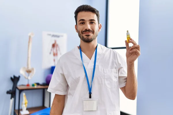 Young Hispanic Physiotherapist Man Holding Cbd Oil Pain Relief Looking — Stockfoto