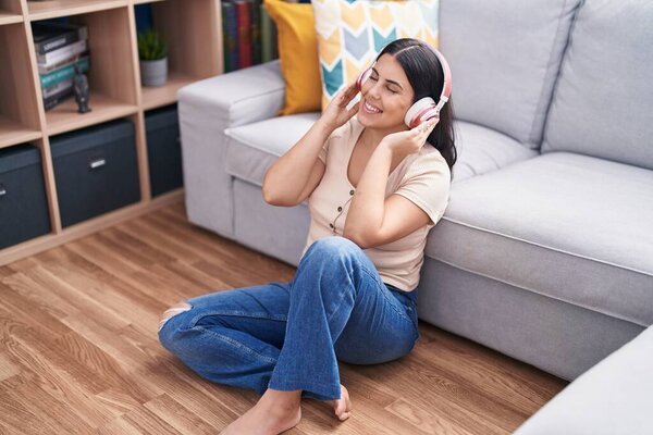 Young beautiful hispanic woman listening to music sitting on floor at home