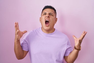 Young hispanic man standing over pink background crazy and mad shouting and yelling with aggressive expression and arms raised. frustration concept. 