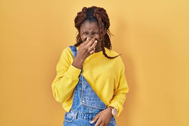 African woman standing over yellow background smelling something stinky and disgusting, intolerable smell, holding breath with fingers on nose. bad smell 