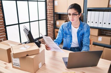 Young beautiful hispanic woman ecommerce using laptop reading document at office