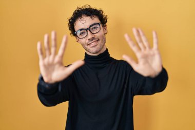 Hispanic man standing over yellow background showing and pointing up with fingers number ten while smiling confident and happy. 