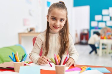 Adorable hispanic girl preschool student sitting on table drawing on paper at kindergarten