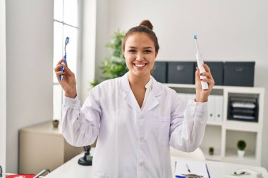 Young hispanic dentist woman holding ordinary toothbrush and electric toothbrush smiling with a happy and cool smile on face. showing teeth. 