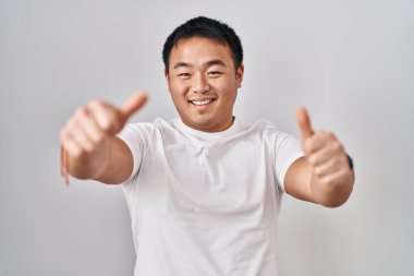 Young chinese man standing over white background approving doing positive gesture with hand, thumbs up smiling and happy for success. winner gesture. 
