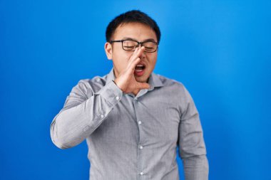 Young chinese man standing over blue background shouting and screaming loud to side with hand on mouth. communication concept. 