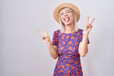 Young caucasian woman wearing flowers dress and summer hat smiling with tongue out showing fingers of both hands doing victory sign. number two. 