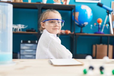 Sevimli sarışın kız öğrenci kendinden emin gülümsüyor. Laboratuvar sınıfında kollarını kavuşturup el kol hareketi yapıyor.