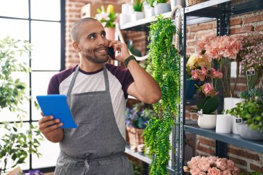 Young latin man florist talking on smartphone using touchpad at flower shop