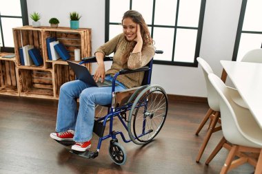Young beautiful hispanic woman using laptop sitting on wheelchair at home