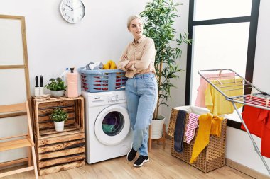 Genç beyaz kadın gülümsüyor. Çamaşırhanede kollarını kavuşturmuş duruyor.