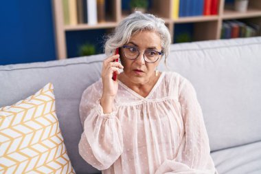 Orta yaşlı, kır saçlı, akıllı telefondan konuşan, endişeli yüzlü bir kadın.
