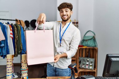 Young arab man smiling confident holding shopping bag working at clothing store