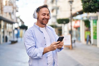 Genç, beyaz bir adam gülerek sokakta müzik dinliyor.
