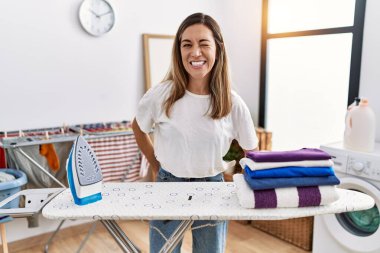 Young hispanic woman ironing clothes at laundry room winking looking at the camera with sexy expression, cheerful and happy face. 