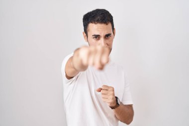 Handsome hispanic man standing over white background punching fist to fight, aggressive and angry attack, threat and violence 