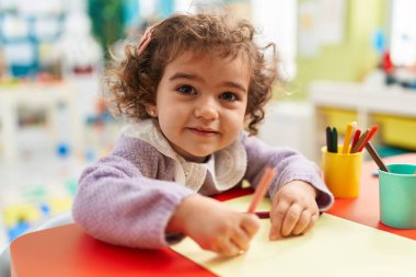 Adorable hispanic girl preschool student sitting on table drawing on paper at kindergarten
