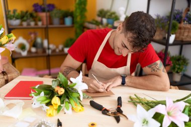 Young hispanic man florist smiling confident writing on envelope letter at flower shop