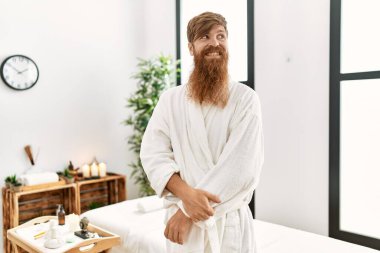 Redhead man with long beard wearing bathrobe at wellness spa looking away to side with smile on face, natural expression. laughing confident. 