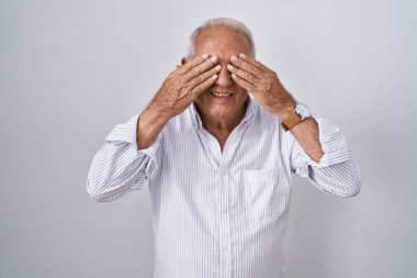 Senior man with grey hair standing over isolated background covering eyes with hands smiling cheerful and funny. blind concept. 
