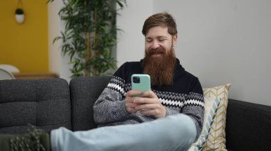 Young redhead man using smartphone sitting on sofa at home