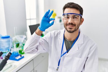 Young hispanic man wearing scientist uniform holding pill at laboratory