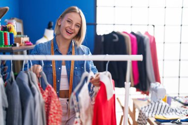Young blonde woman tailor smiling confident holding clothes on rack at sewing studio