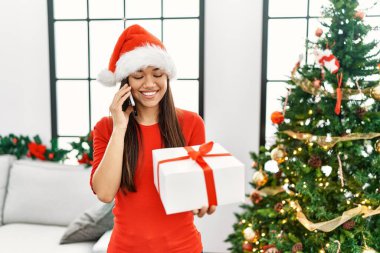 Genç Latin kadın akıllı telefonda konuşuyor. Yılbaşı ağacının yanında duruyor.