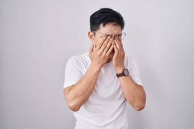Young asian man standing over white background rubbing eyes for fatigue and headache, sleepy and tired expression. vision problem 