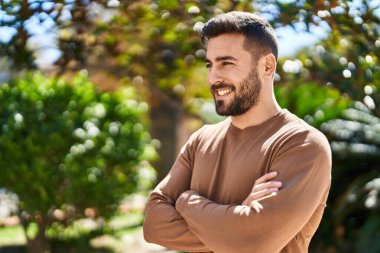 Genç İspanyol adam gülümsüyor. Parkta kollarını kavuşturmuş duruyor.