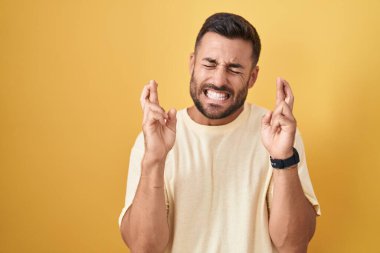Handsome hispanic man standing over yellow background gesturing finger crossed smiling with hope and eyes closed. luck and superstitious concept. 