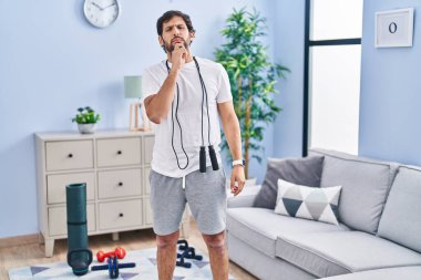 Handsome latin man wearing sportswear at home looking confident at the camera with smile with crossed arms and hand raised on chin. thinking positive. 