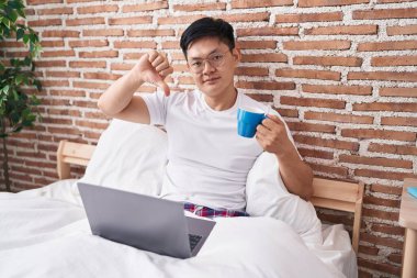 Young asian man drinking coffee sitting on the bed with angry face, negative sign showing dislike with thumbs down, rejection concept 