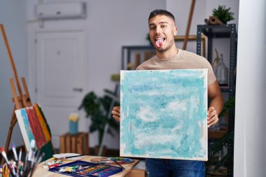 Young hispanic painter man holding canvas sticking tongue out happy with funny expression. 
