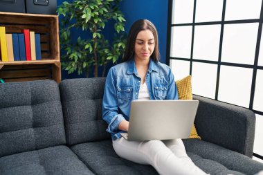 Hispanic woman using laptop at home thinking attitude and sober expression looking self confident 