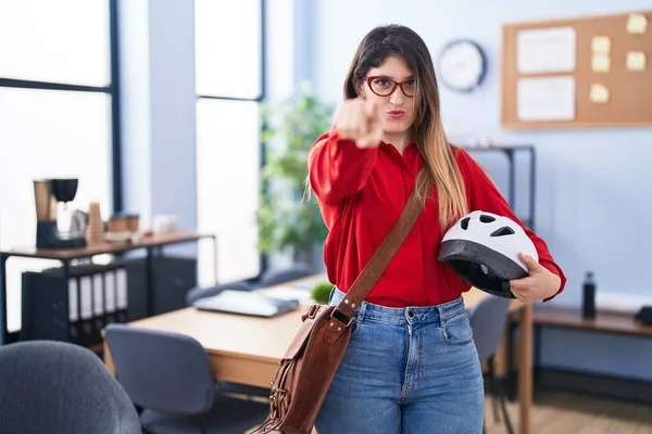 Young brunette woman working at the office holding bike helmet pointing with finger to the camera and to you, confident gesture looking serious